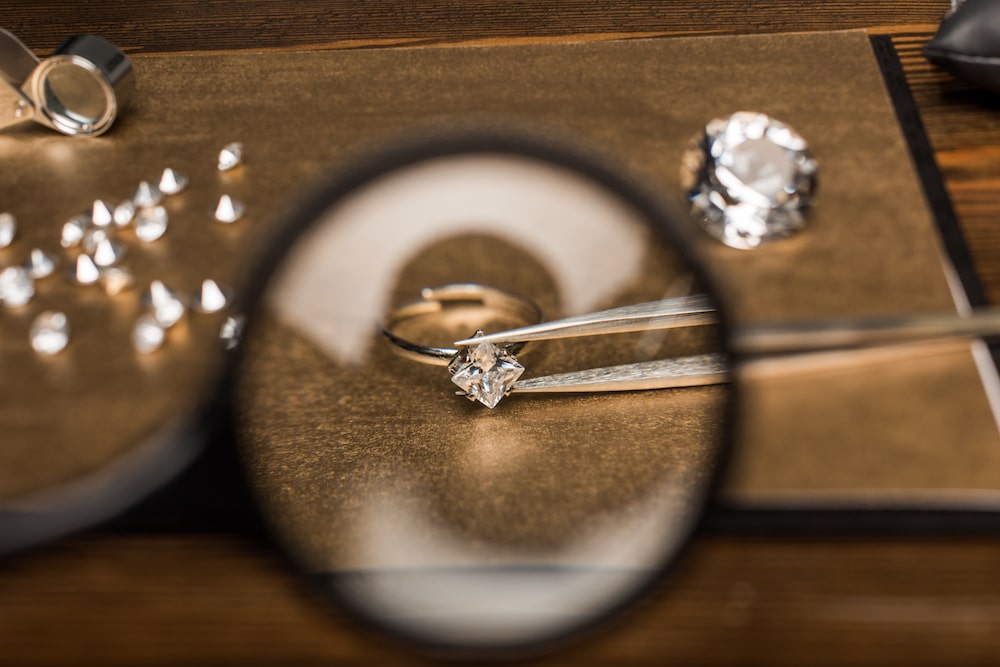 Selective focus of magnifying glass, jewelry ring with gemstone in tweezers on board on wooden table