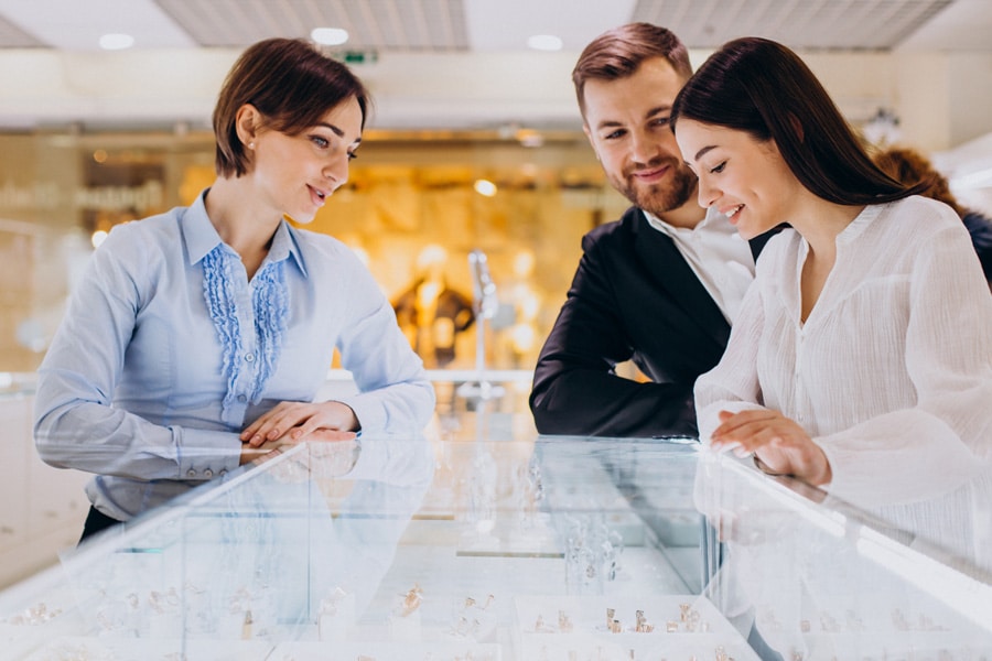 A couple looks into a jewelry display case and talks with a jeweler while they shop for jewelry at a store