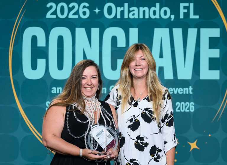 Two women posing together, one holding a glass award, in front of a Conclave 2026 Orlando, FL backdrop.