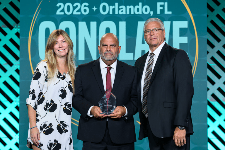 Three people posing together, one holding a glass award, in front of a Conclave 2026 Orlando, FL backdrop.