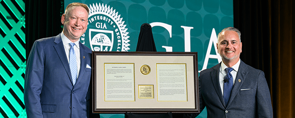 Two men posing on stage with a framed Robert M. Shipley Award certificate in front of a GIA backdrop.