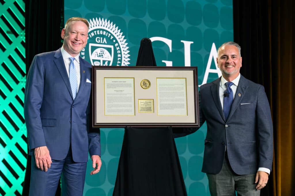 Two men posing on stage with a framed Robert M. Shipley Award certificate in front of a GIA backdrop.